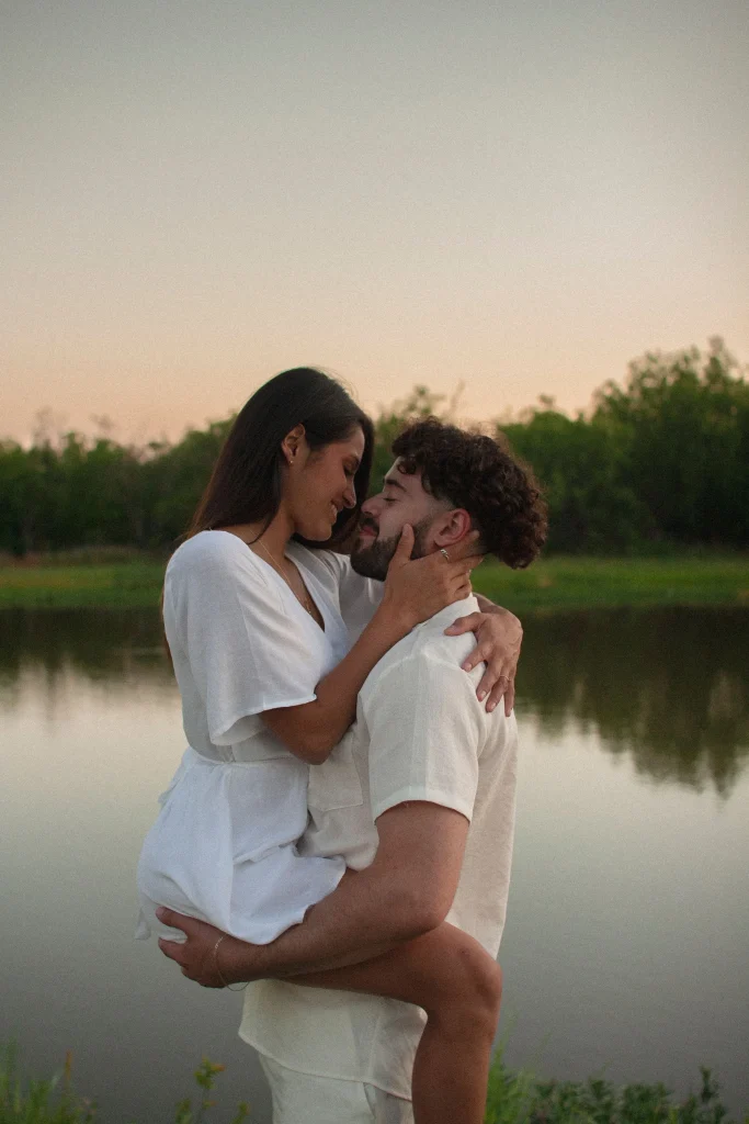 Couple embracing by a lake, the woman lifted in the man’s arms as they smile at each other.
