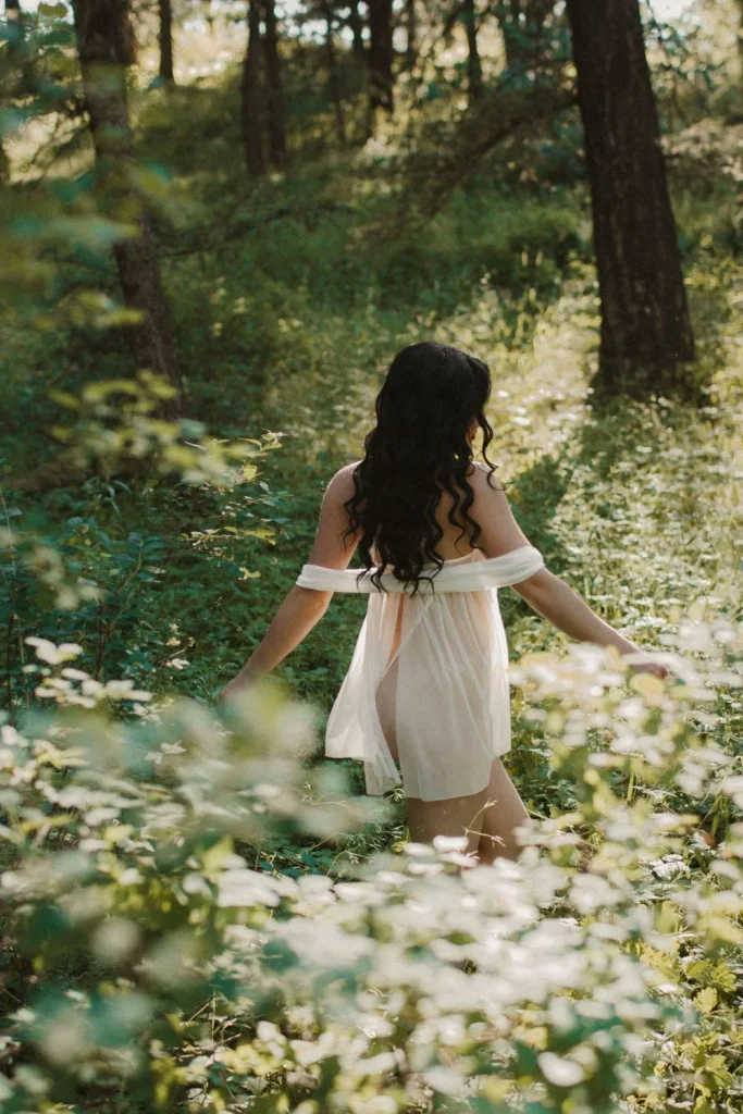 Back view of a woman walking through a lush forest in a sheer white dress, surrounded by tall plants and dappled sunlight.