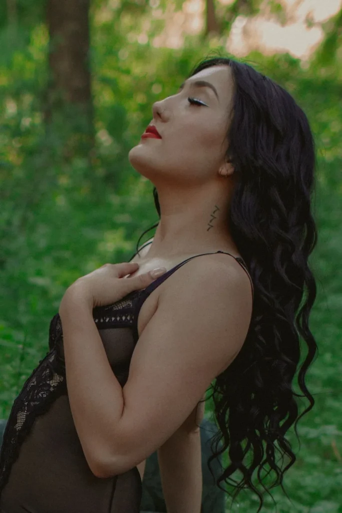 Close-up of a woman in a sheer black bodysuit in a forest, head tilted back with eyes closed, hand resting on her chest.