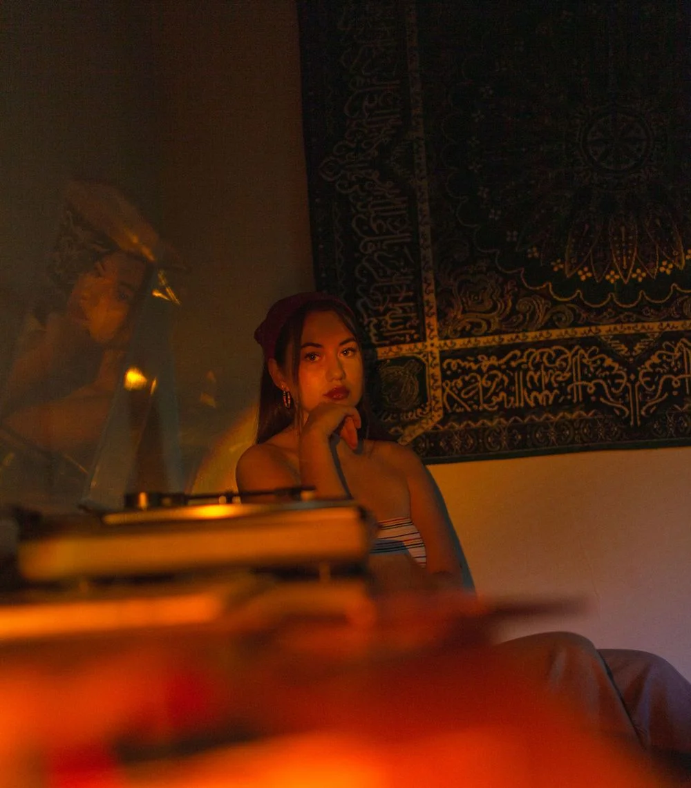 (Duplicate of previous image) Woman sitting in warm, low lighting beside a turntable, her face softly illuminated against a patterned tapestry.