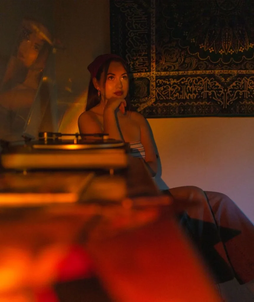 Woman sitting near a record player in atmospheric golden light, gazing upward with a reflective expression, tapestry backdrop behind her.