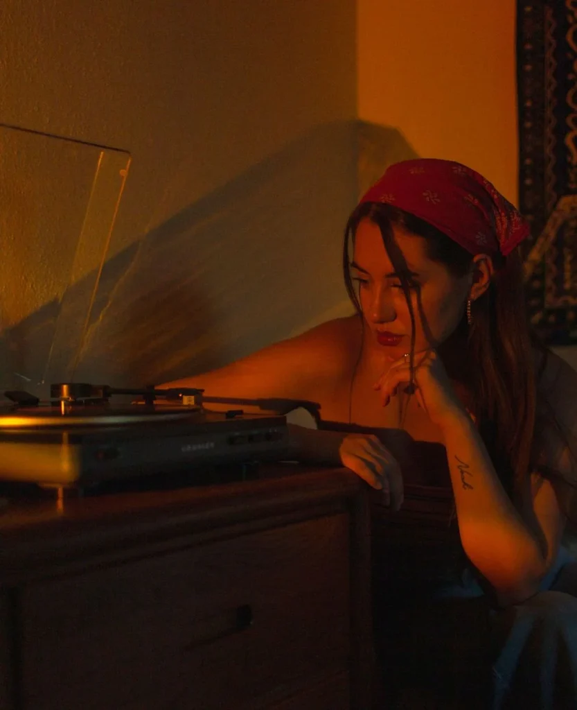 Woman adjusting a record player in warm, moody lighting, wearing a red bandana and resting her chin on her hand.