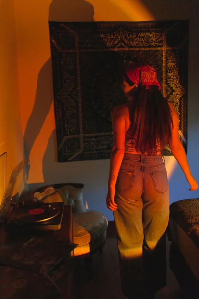 Woman standing in a dimly lit room facing a decorative tapestry, with warm orange light casting her shadow on the wall.