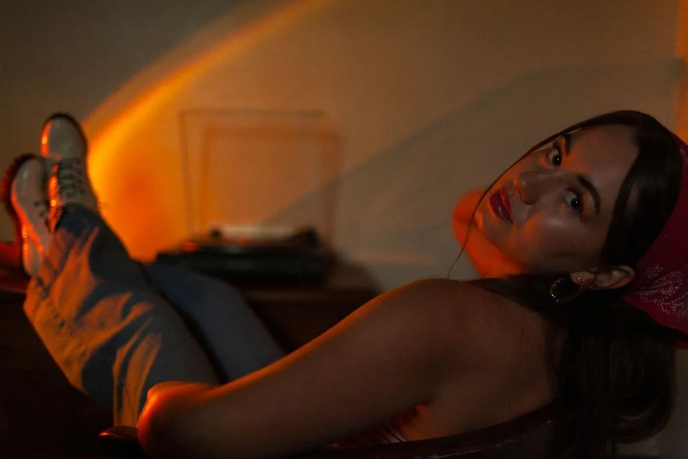 Woman reclining in a chair with her feet up near a record player, looking back over her shoulder in soft orange light.