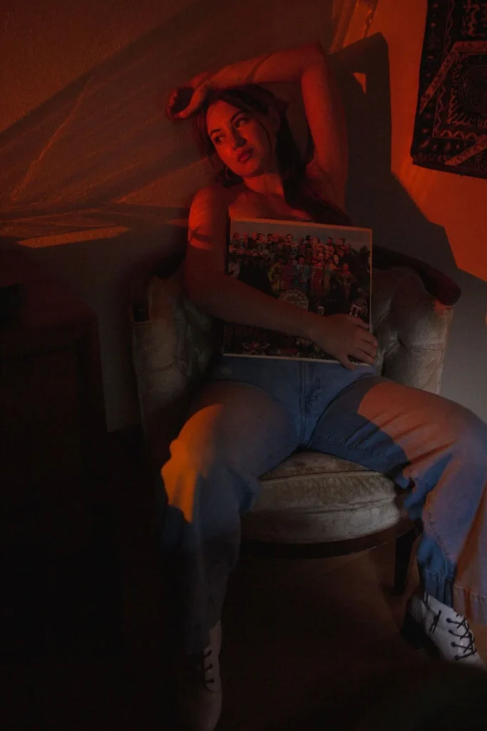 Woman sitting in a chair holding a vinyl record against her chest, bathed in dramatic red and orange lighting.