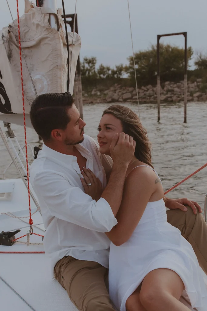 Man gently touching a woman’s face as they sit together on a sailboat with water in the background.