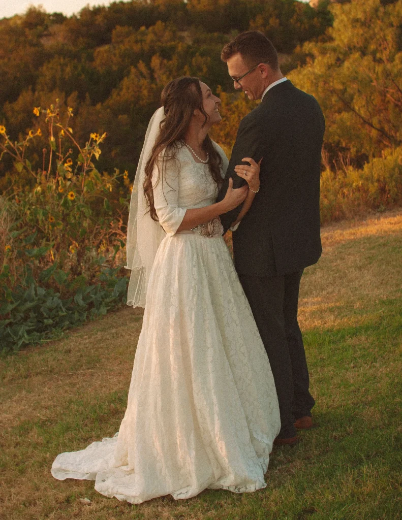 Bride and groom standing together smiling in a grassy field during golden hour.