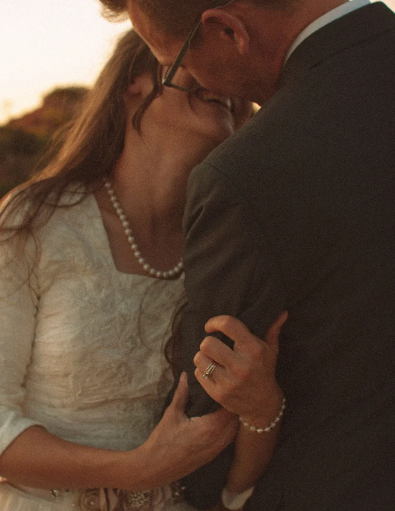 Close-up of bride holding groom’s arm as they lean in with soft smiles at sunset.