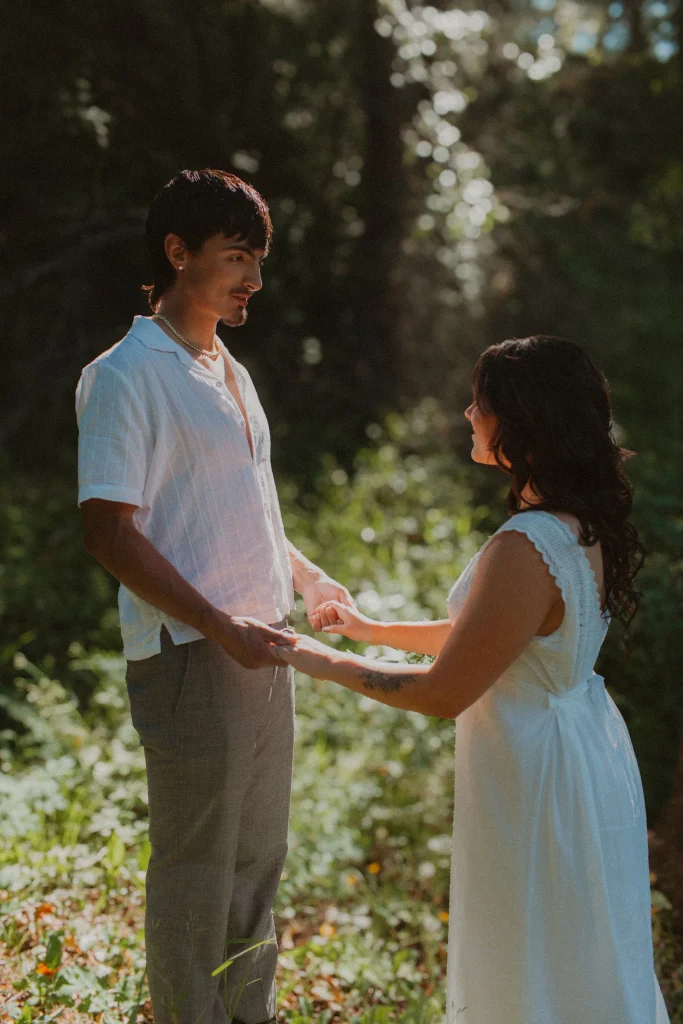 Couple standing in a sunlit forest clearing, holding hands and looking at each other.