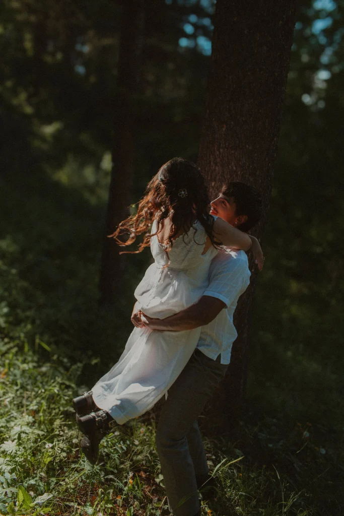 Man lifts woman in a white dress in a sunlit forest clearing, both smiling and embraced in motion.
