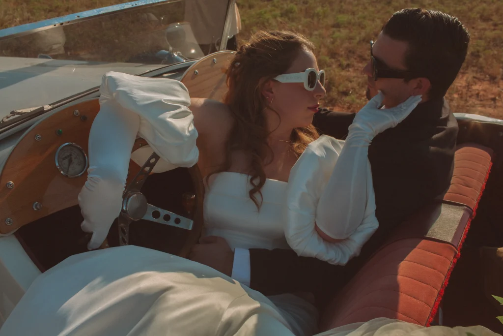 Bride in statement sleeves and sunglasses leaning toward the groom in a vintage convertible, touching his face.