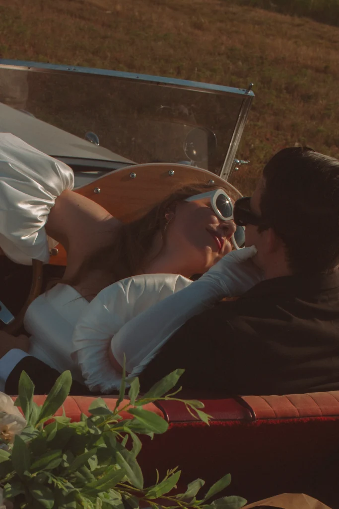 Bride reaching toward groom for a kiss while reclining across the front seat of a classic convertible.