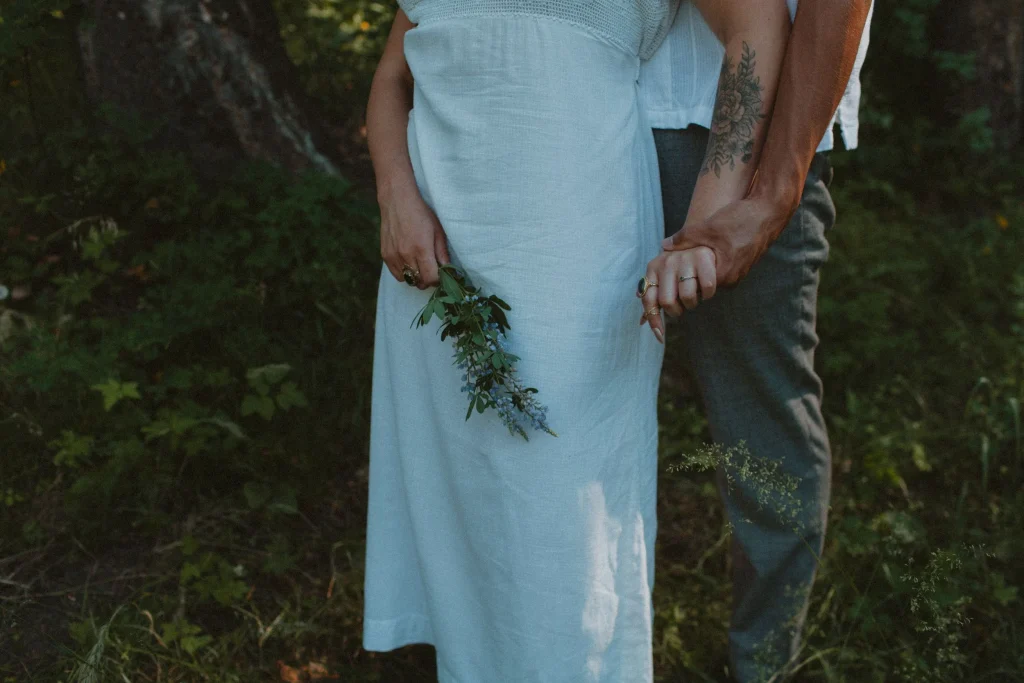 Couple standing close together in the forest holding hands, woman holding a small bouquet of wildflowers.