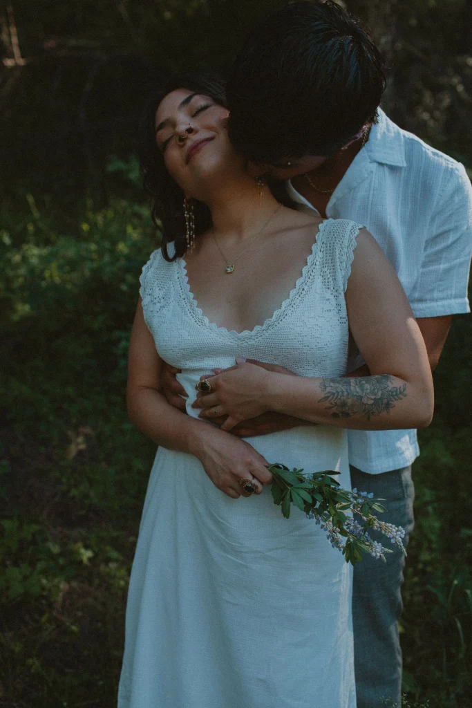 Woman in a white dress smiles softly as her partner hugs her from behind and kisses her neck in a forest.