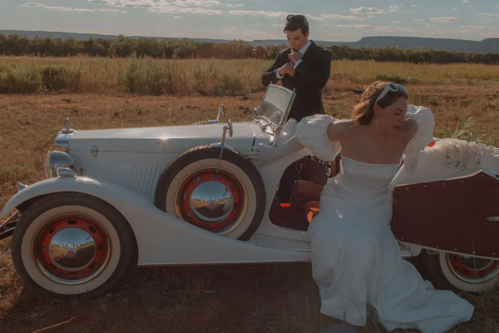 Groom adjusting his cufflinks beside a vintage car as the bride steps out in her wedding gown.