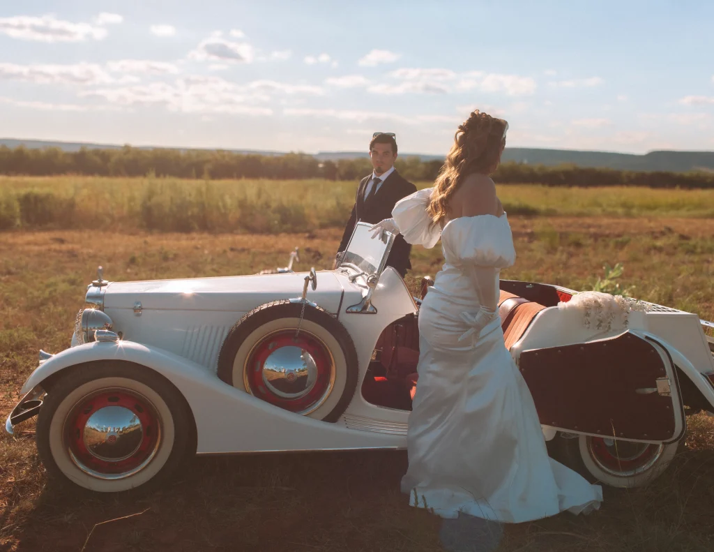 Groom adjusting his cufflinks beside a vintage car as the bride steps out in her wedding gown.