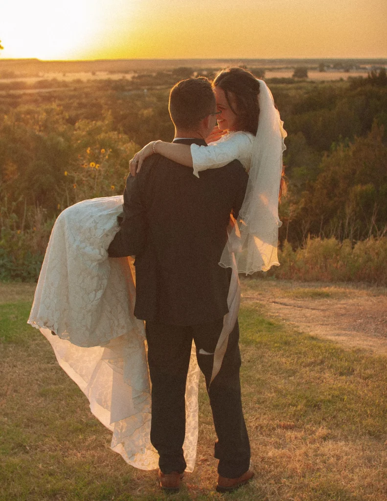 Bride standing beside a vintage white convertible with the groom in the background, golden hour light across the field.