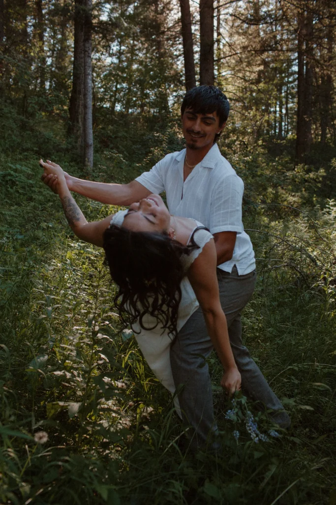 Man playfully dips woman backward while dancing in a sunlit forest meadow.