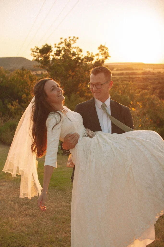 Groom holding bride in his arms at sunset as she laughs, her veil glowing in golden light.
