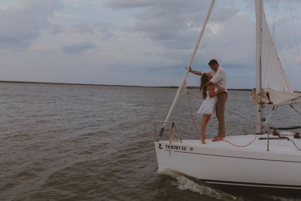 Couple stands at the bow of a sailboat, embracing as the boat moves across the water under cloudy skies.