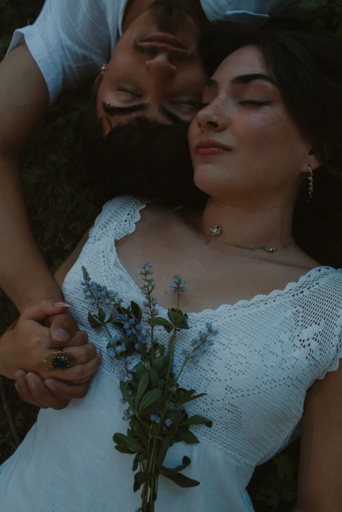 Close-up of couple lying side by side in the forest holding hands, wildflowers resting on the woman's dress.