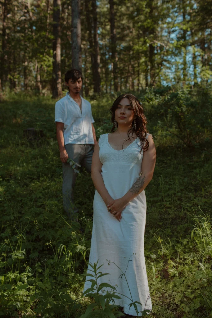 Woman in white dress stands in a sunlit forest while her partner stands softly blurred behind her holding flowers.