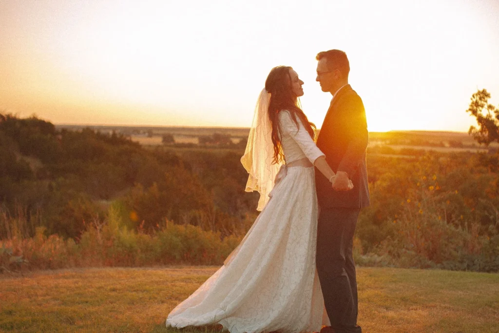 Bride and groom holding hands at sunset, smiling at each other as her lace gown flows behind her.