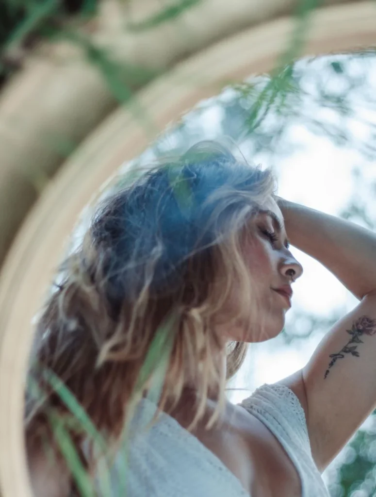 Reflection of a woman in a circular mirror outdoors, hand in her hair, surrounded by greenery.