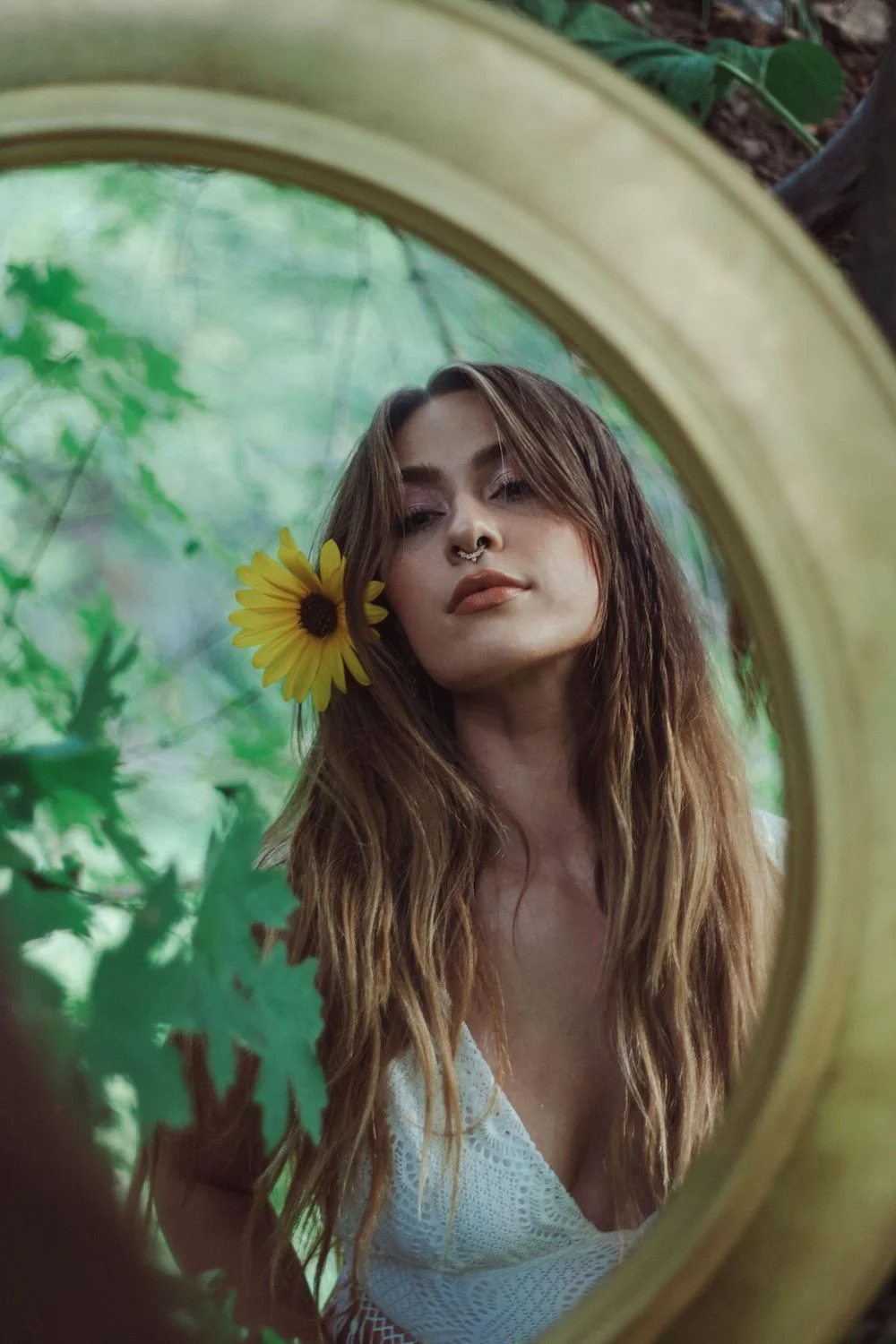 Mirror reflection of a woman with a yellow flower tucked behind her ear, framed by forest greenery.