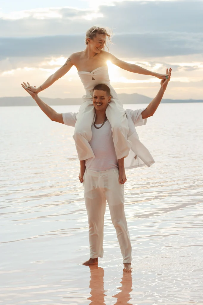 Woman sitting on partner’s shoulders with arms outstretched over reflective water at sunset.