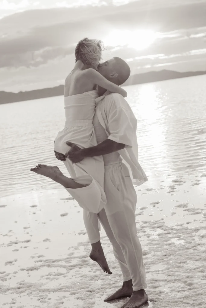 Black-and-white image of a couple embracing and kissing while standing barefoot on a salt flat.